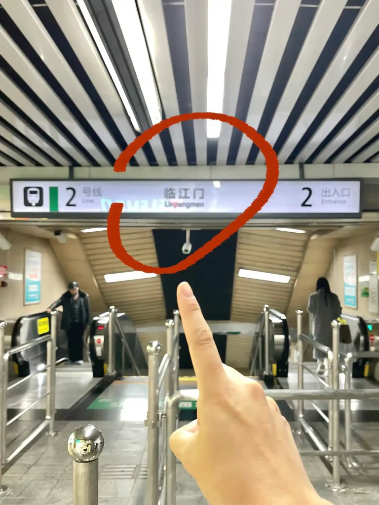 A hand points to the Linjiangmen metro station sign circled in red above the escalators, with Exit 2 marked on the right